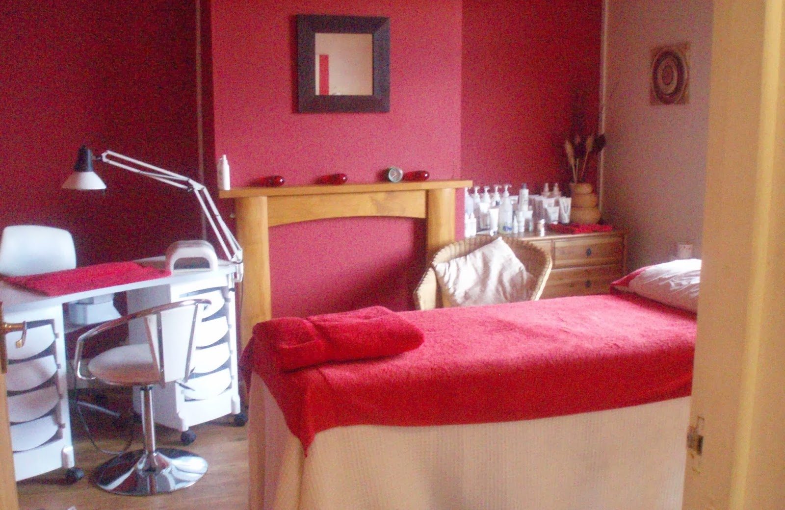 A treatment room at Pastiche: deep red walls, a couch made up with red towels, a pine mantelpiece and a shelf of skincare